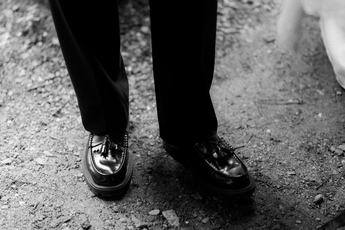 Feet in black trousers and polished shoes on rough ground.