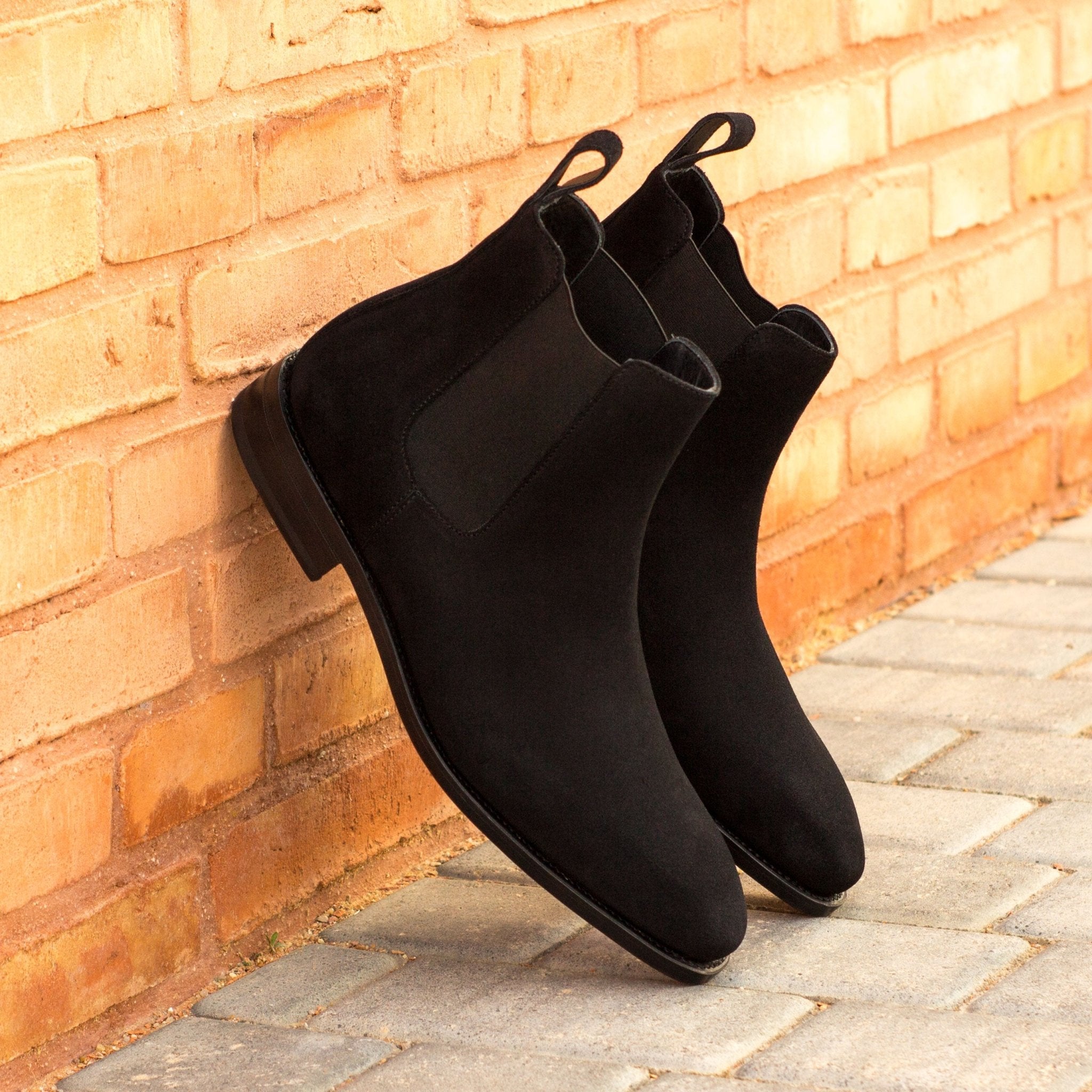 Black Chelsea boots leaning against a brick wall on a stone surface.