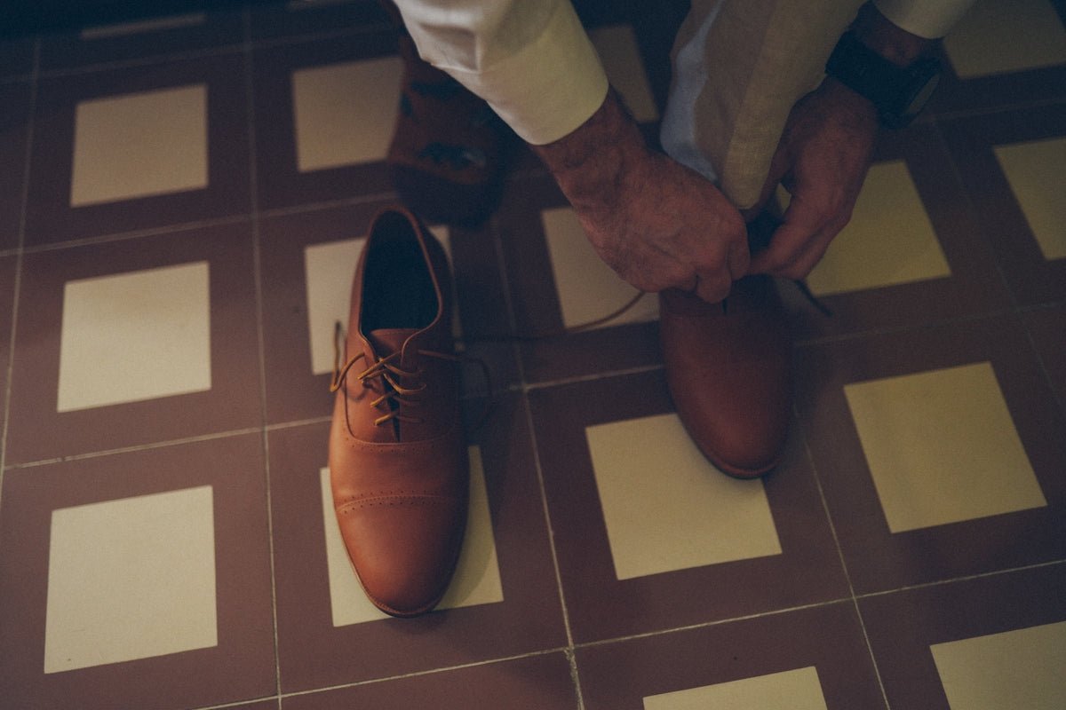 A person tying brown leather dress shoes on tiled floor.