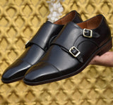 A pair of black double monk shoes with a hand-stitched border sole, held up against a yellow background.