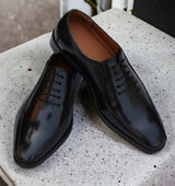 A pair of black, handmade leather shoes with a lace-up closure, displayed on a textured surface.